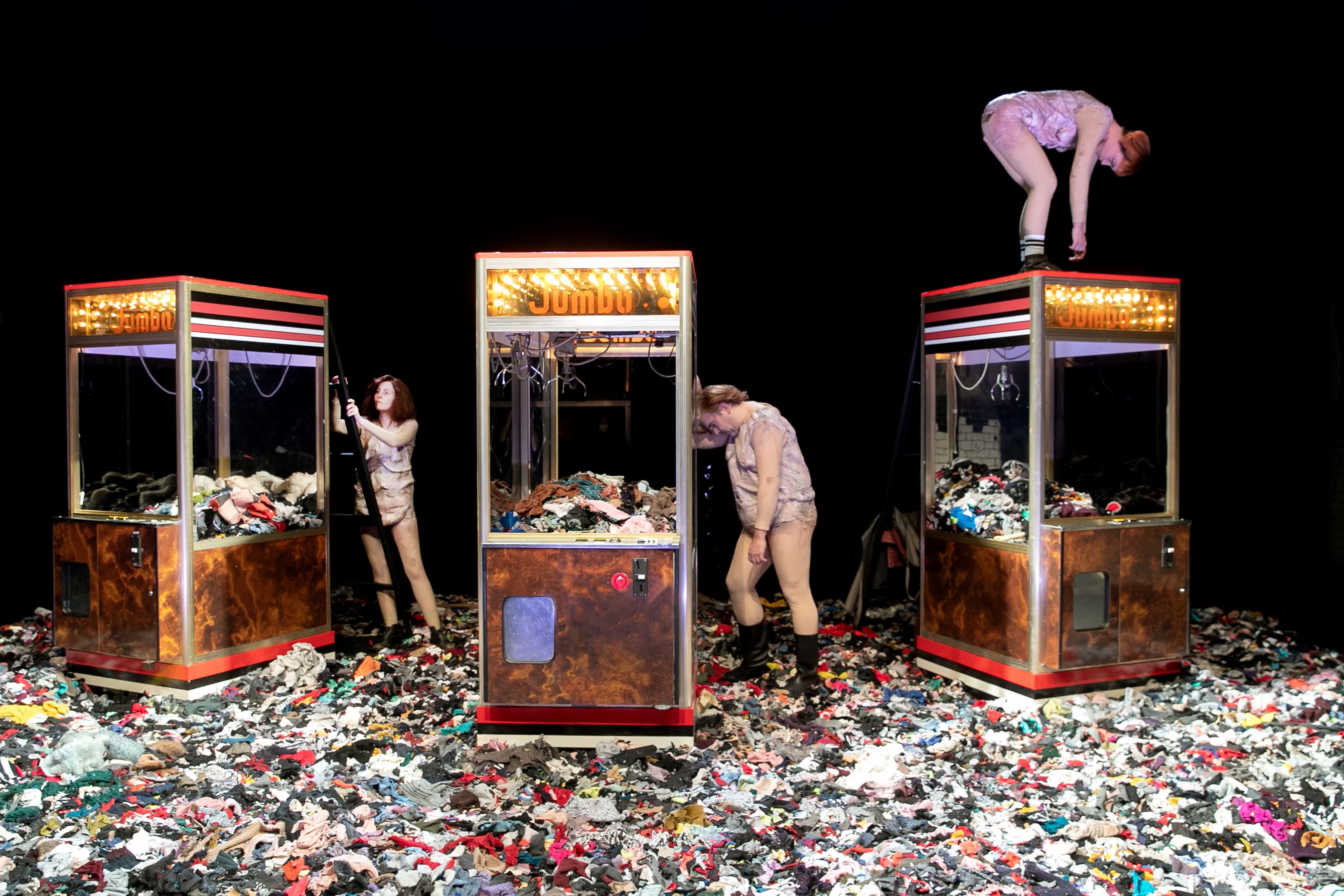 Szenenfoto aus Die Weber:innen: Spielautomaten und Darsteller:innen auf einer Bühne voller Stoffetzen